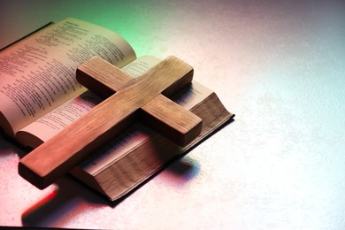 Photo of Wooden cross and Bible on textured table in color lights, closeup with space for text. Religion of Christianity