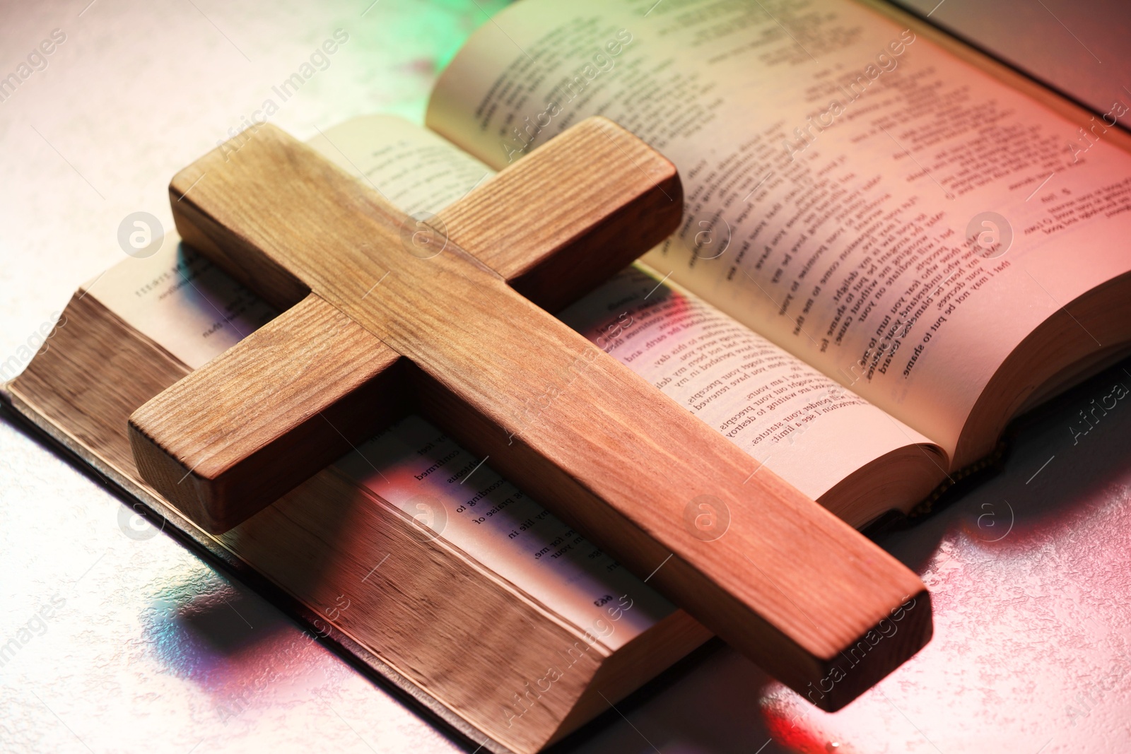 Photo of Wooden cross and Bible on textured table in color lights, closeup. Religion of Christianity