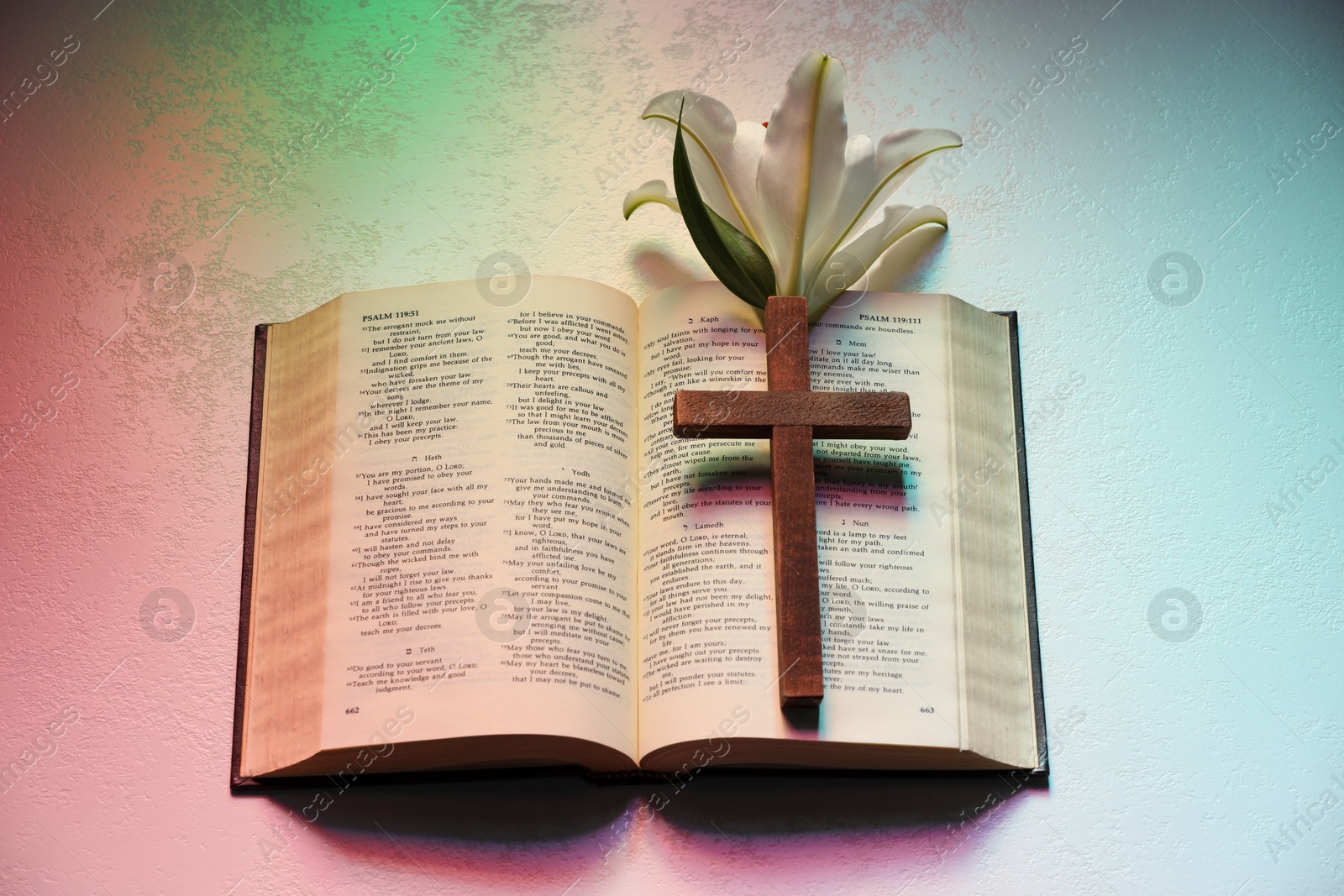 Photo of Wooden cross, Bible and lily flower on textured table in color lights, top view. Religion of Christianity