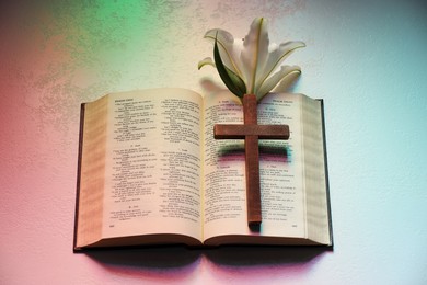 Wooden cross, Bible and lily flower on textured table in color lights, top view. Religion of Christianity