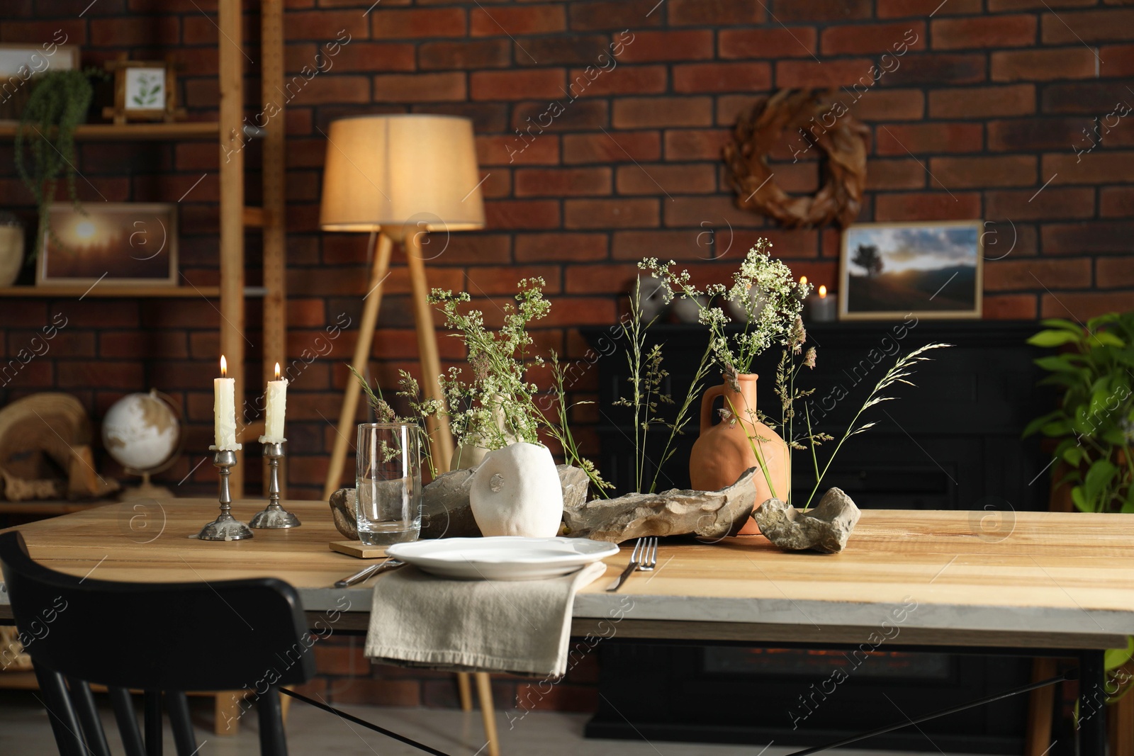 Photo of Set of clean dishware, vases with beautiful flowers, stones and burning candles on wooden table in stylish dining room