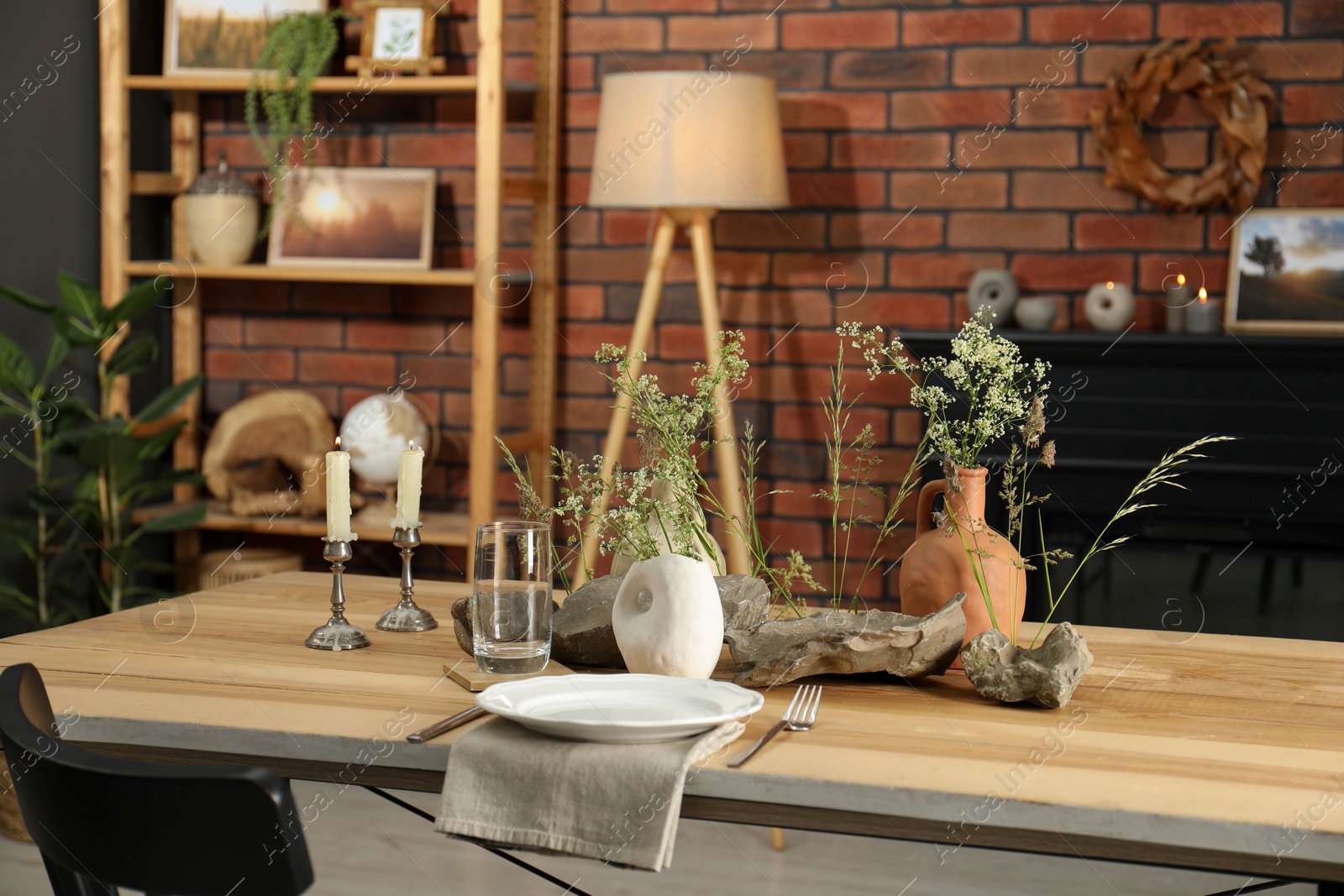 Photo of Set of clean dishware, vases with beautiful flowers, stones and burning candles on wooden table in stylish dining room