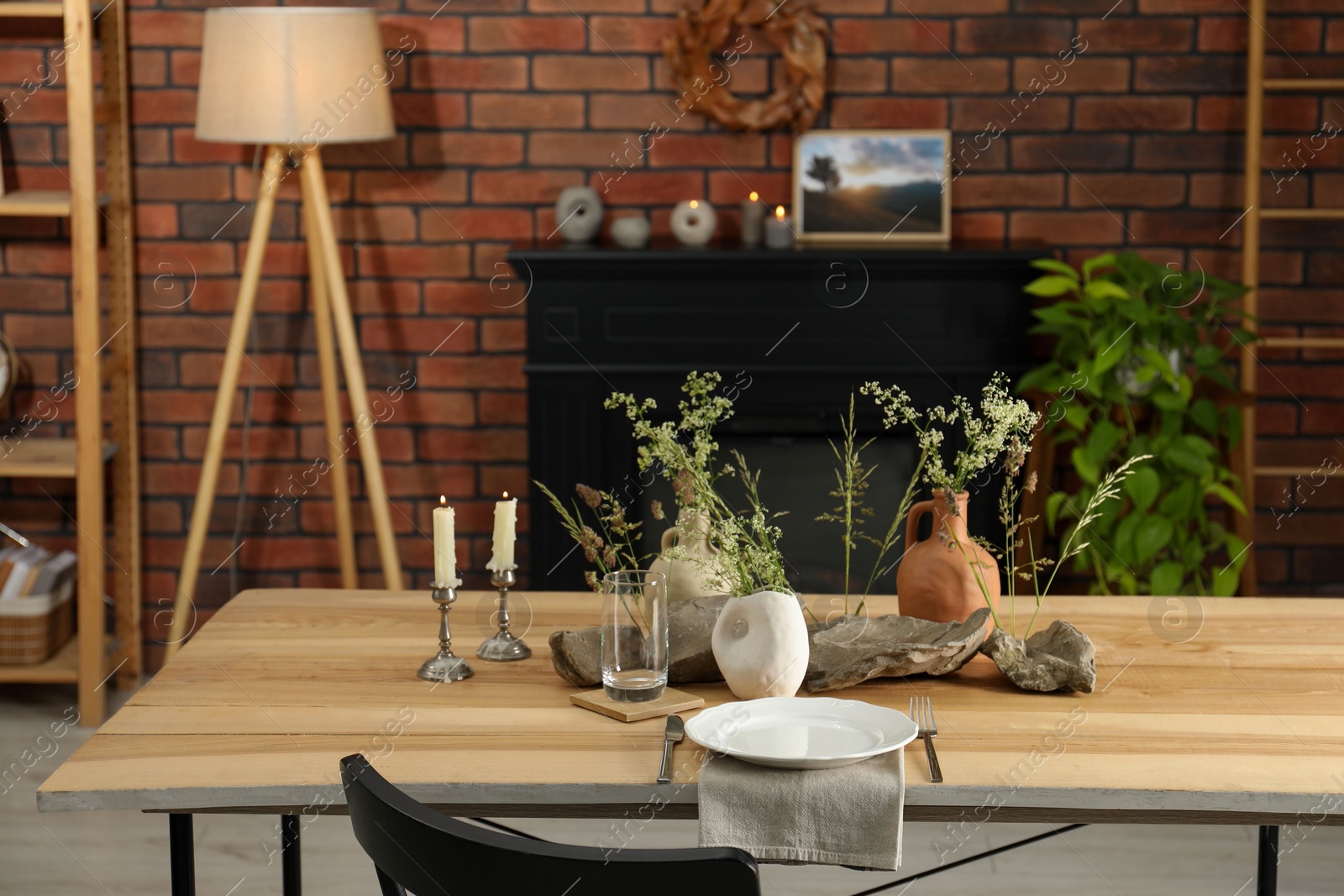 Photo of Set of clean dishware, vases with beautiful flowers, stones and burning candles on wooden table in stylish dining room