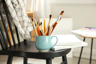 Photo of Canvases, paints and cup of brushes on chair in workshop