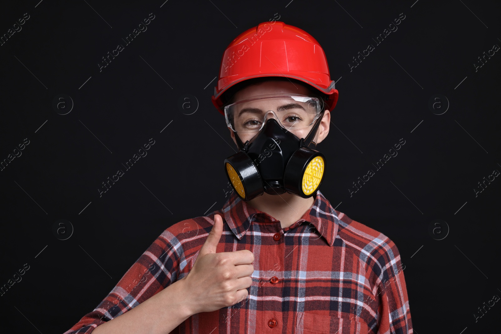 Photo of Woman in respirator, protective glasses and helmet showing thumbs up on black background