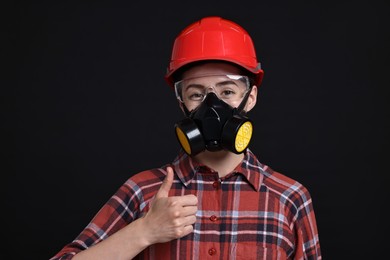 Photo of Woman in respirator, protective glasses and helmet showing thumbs up on black background