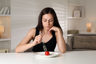 Eating disorder. Sad woman holding fork with tomato at table indoors