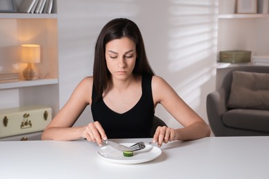 Eating disorder. Sad woman cutting cucumber at white table indoors