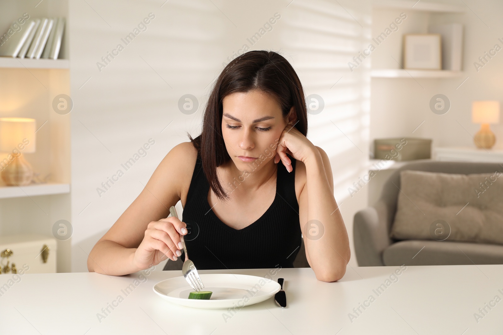 Photo of Eating disorder. Sad woman holding fork with cucumber at white table indoors