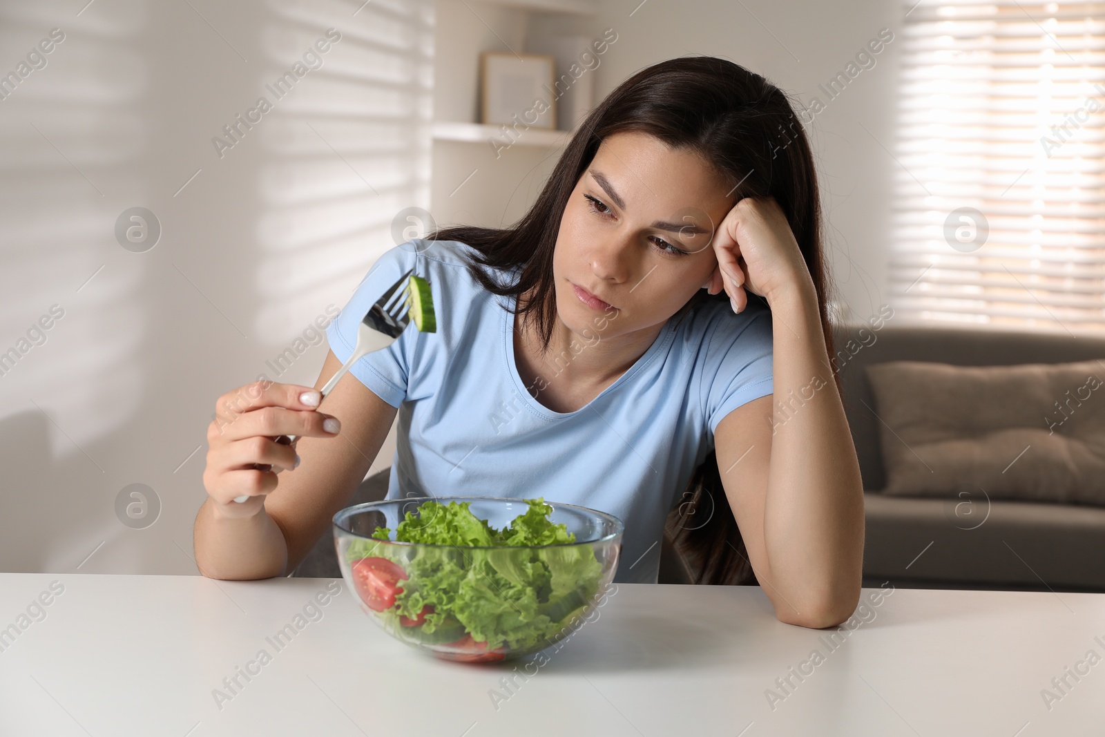 Photo of Eating disorder. Sad woman holding fork with cucumber over bowl at white table indoors