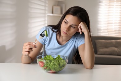 Eating disorder. Sad woman holding fork with cucumber over bowl at white table indoors