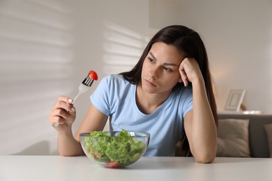 Eating disorder. Sad woman holding fork with tomato over bowl at white table indoors