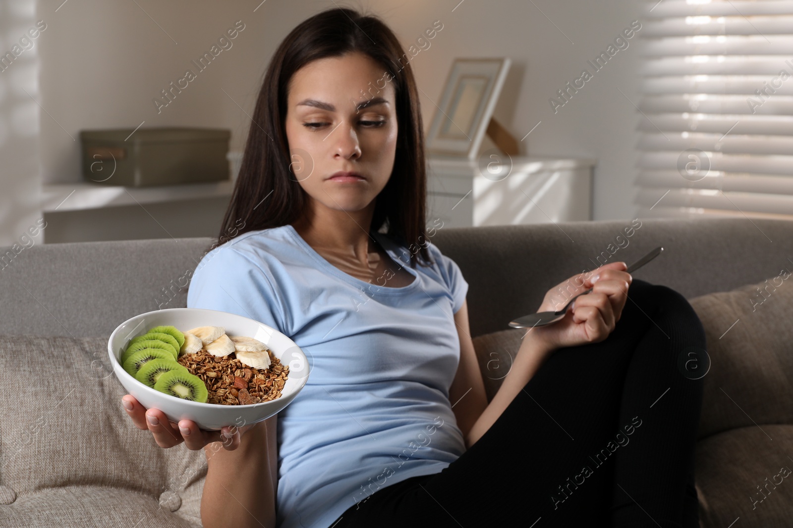 Photo of Eating disorder. Sad woman with spoon and healthy meal on sofa indoors