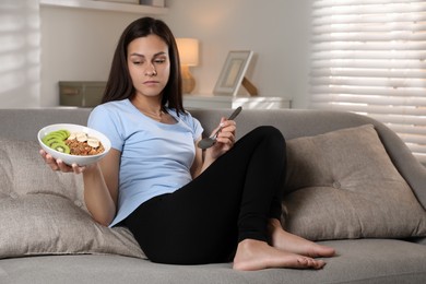 Photo of Eating disorder. Sad woman with spoon and healthy meal on sofa indoors