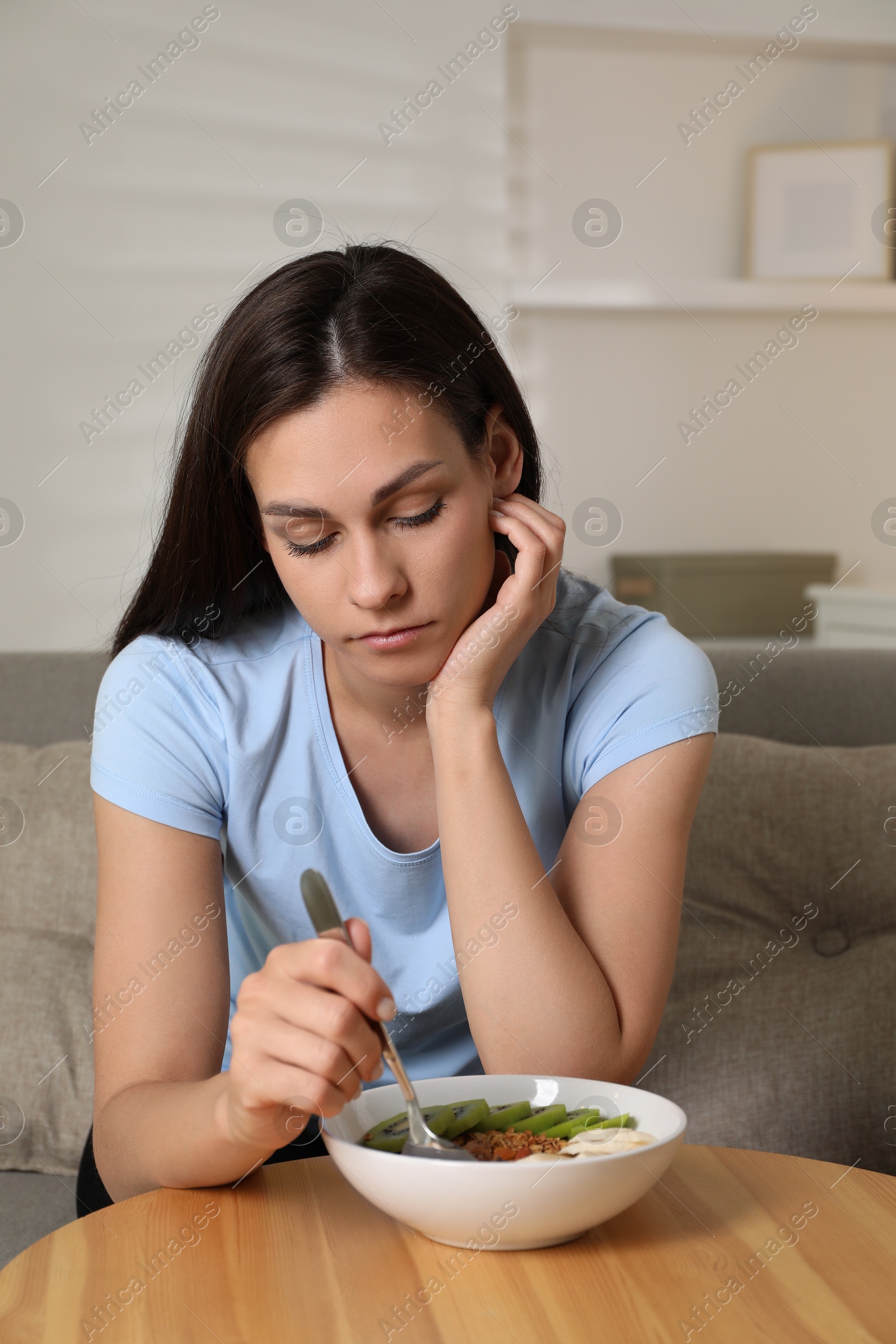 Photo of Eating disorder. Sad woman holding spoon near bowl with granola, kiwi and banana at wooden table indoors