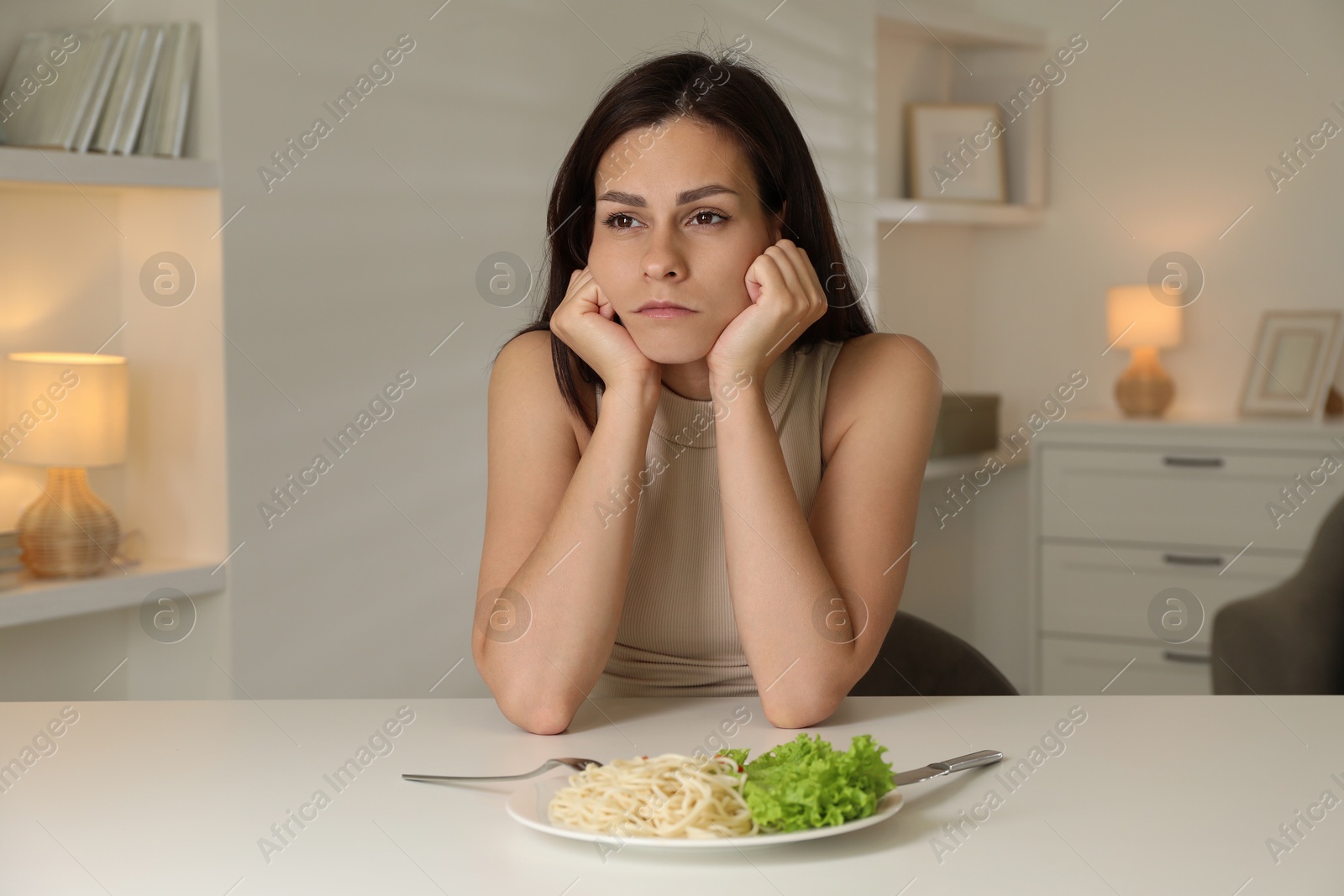 Photo of Eating disorder. Sad woman at white table with spaghetti and cutlery indoors