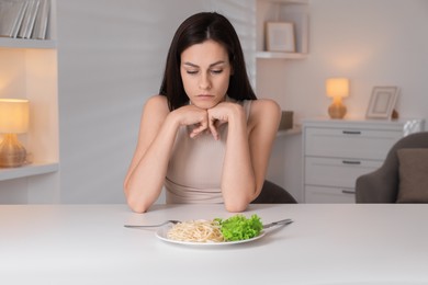 Eating disorder. Sad woman at white table with spaghetti and cutlery indoors