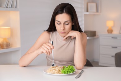 Photo of Eating disorder. Sad woman holding fork near plate with spaghetti at white table indoors