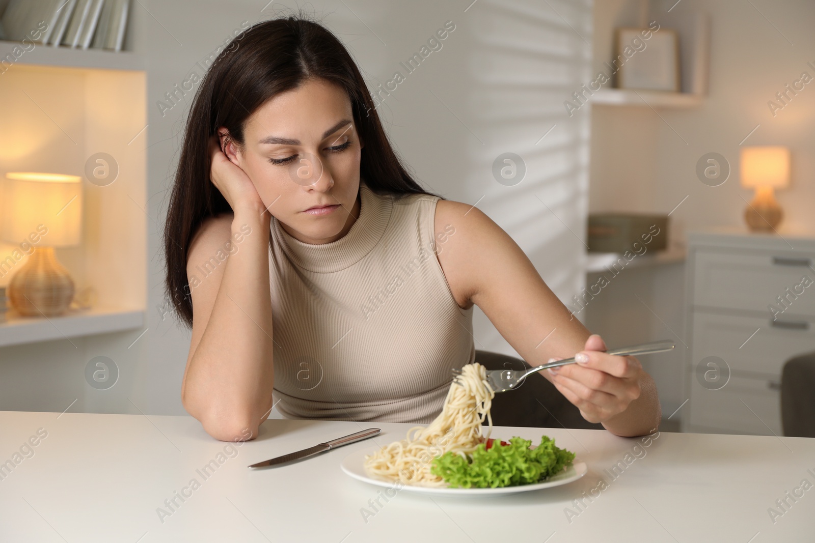 Photo of Eating disorder. Sad woman holding fork with spaghetti over plate at white table indoors