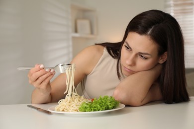 Photo of Eating disorder. Sad woman holding fork with spaghetti over plate at white table indoors