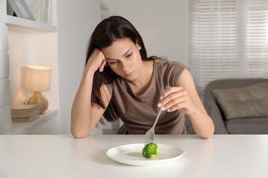 Photo of Eating disorder. Sad woman holding fork with broccoli at white table indoors