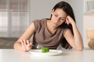 Eating disorder. Sad woman holding fork near broccoli at table indoors