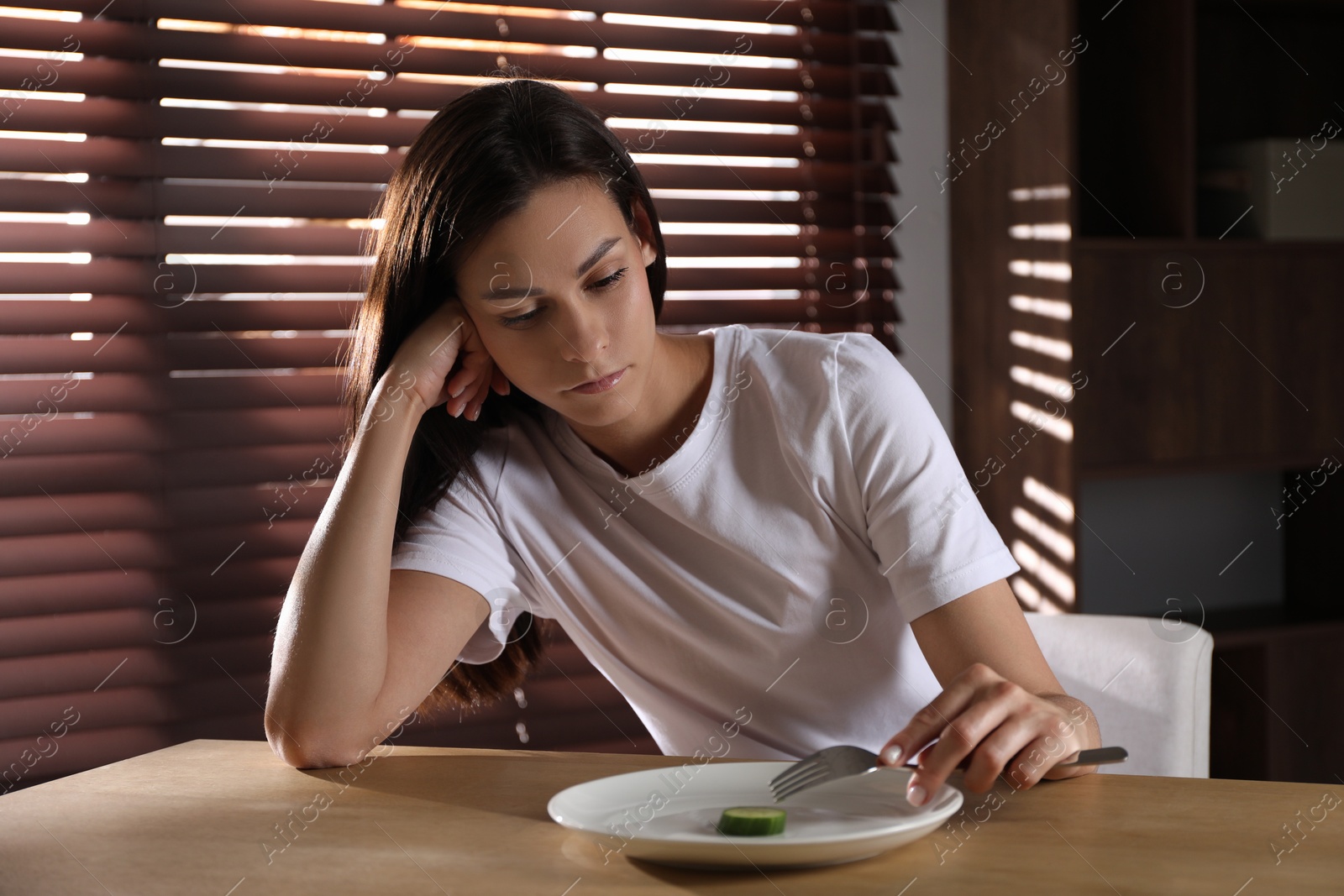 Photo of Eating disorder. Sad woman with fork and slice of cucumber at wooden table