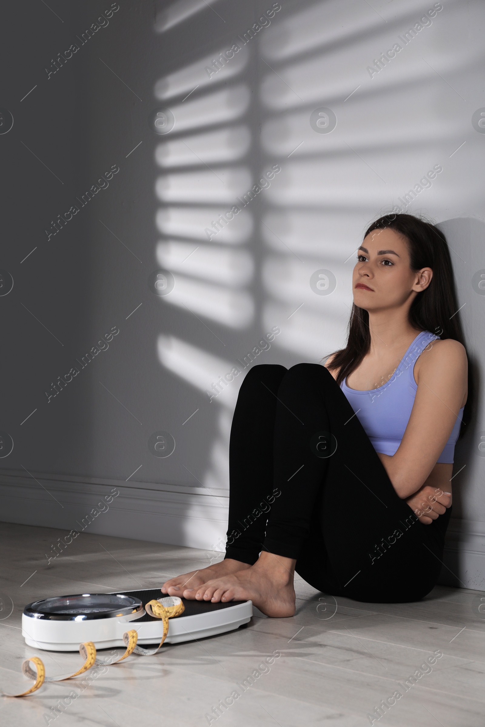 Photo of Eating disorder. Sad woman sitting near scale and measuring tape on floor indoors