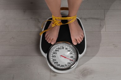 Eating disorder. Woman tied with measuring tape standing on floor scale indoors, top view