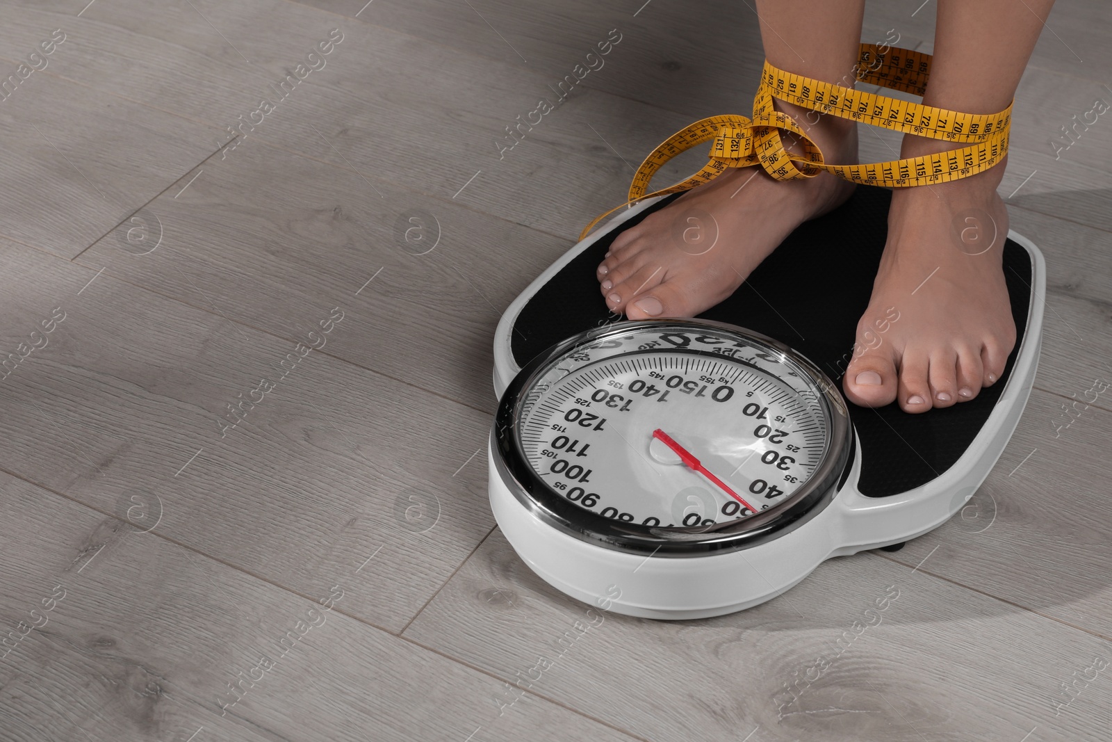 Photo of Eating disorder. Woman tied with measuring tape standing on floor scale indoors, closeup. Space for text