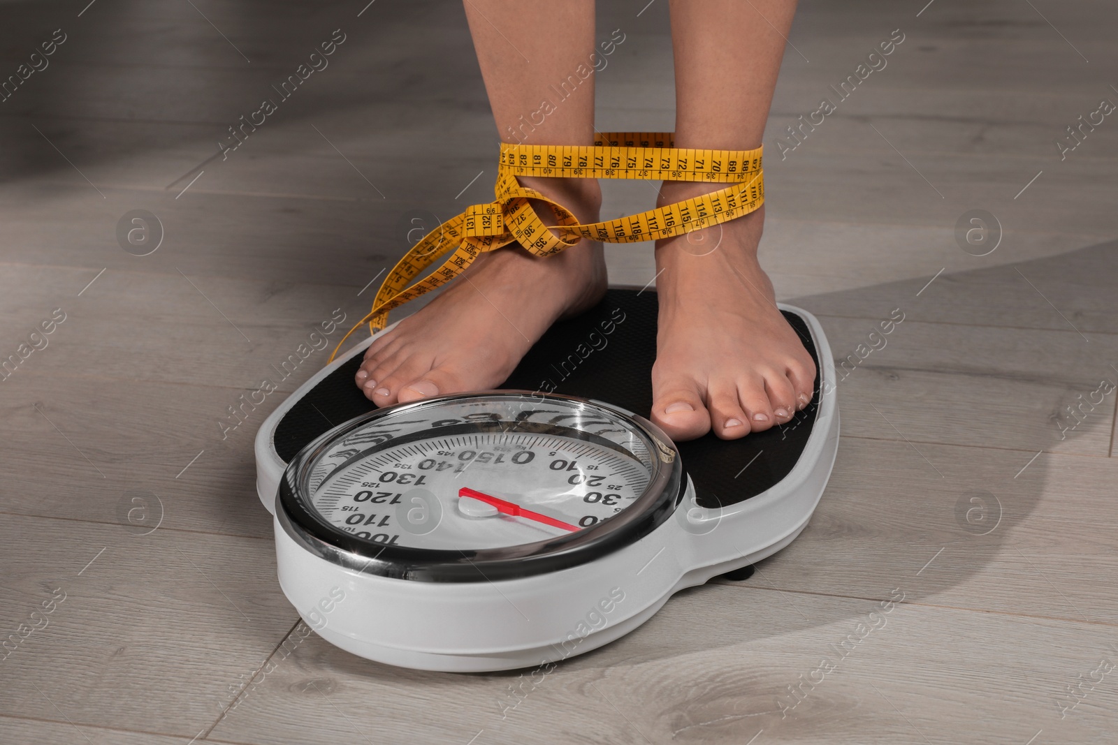 Photo of Eating disorder. Woman tied with measuring tape standing on floor scale indoors, closeup