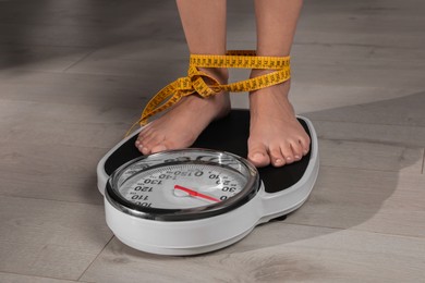 Photo of Eating disorder. Woman tied with measuring tape standing on floor scale indoors, closeup