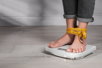 Eating disorder. Woman tied with measuring tape standing on floor scale indoors, closeup. Space for text