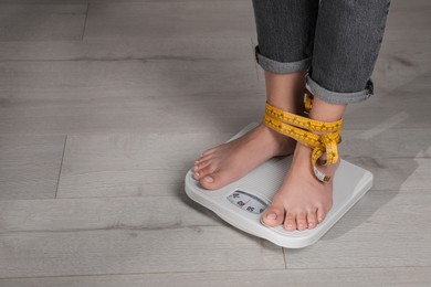 Photo of Eating disorder. Woman tied with measuring tape standing on floor scale indoors, closeup. Space for text