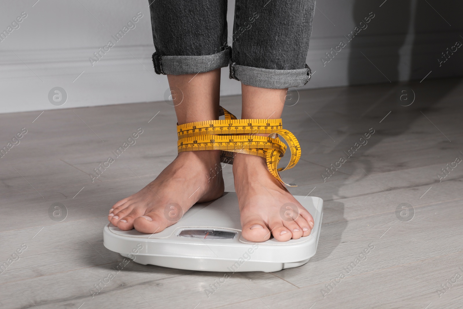 Photo of Eating disorder. Woman tied with measuring tape standing on floor scale indoors, closeup