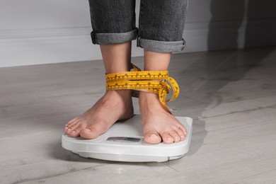 Eating disorder. Woman tied with measuring tape standing on floor scale indoors, closeup