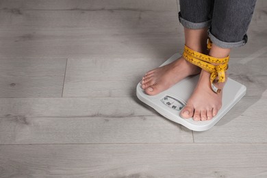 Photo of Eating disorder. Woman tied with measuring tape standing on floor scale indoors, closeup. Space for text