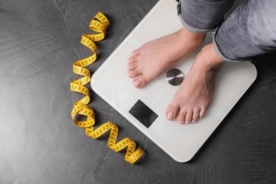 Photo of Eating disorder. Woman standing on floor scale and measuring tape indoors, top view