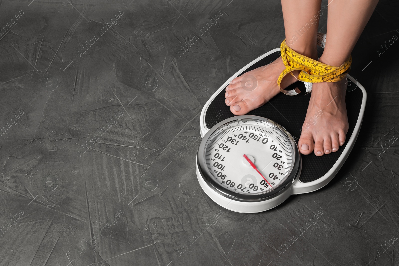 Photo of Eating disorder. Woman tied with measuring tape standing on floor scale indoors, closeup. Space for text