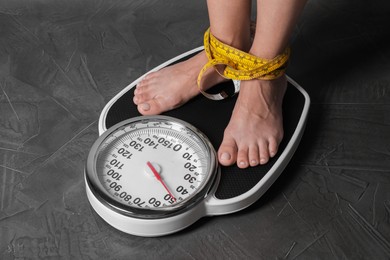 Photo of Eating disorder. Woman tied with measuring tape standing on floor scale indoors, closeup