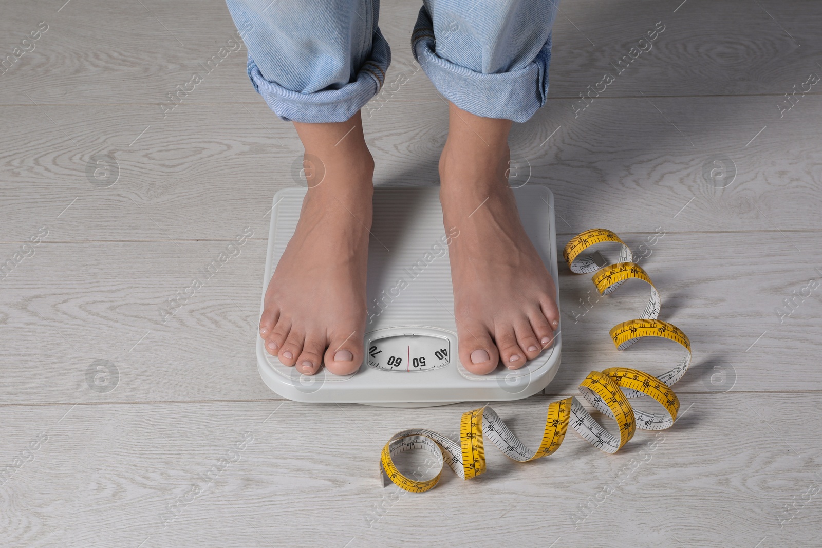 Photo of Eating disorder. Woman standing on floor scale and measuring tape indoors, closeup