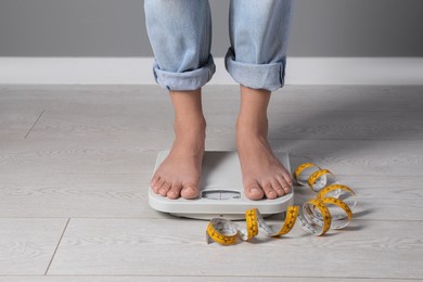 Photo of Eating disorder. Woman standing on floor scale and measuring tape indoors, closeup