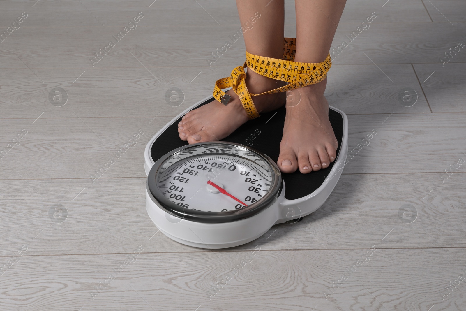 Photo of Eating disorder. Woman tied with measuring tape standing on floor scale indoors, closeup