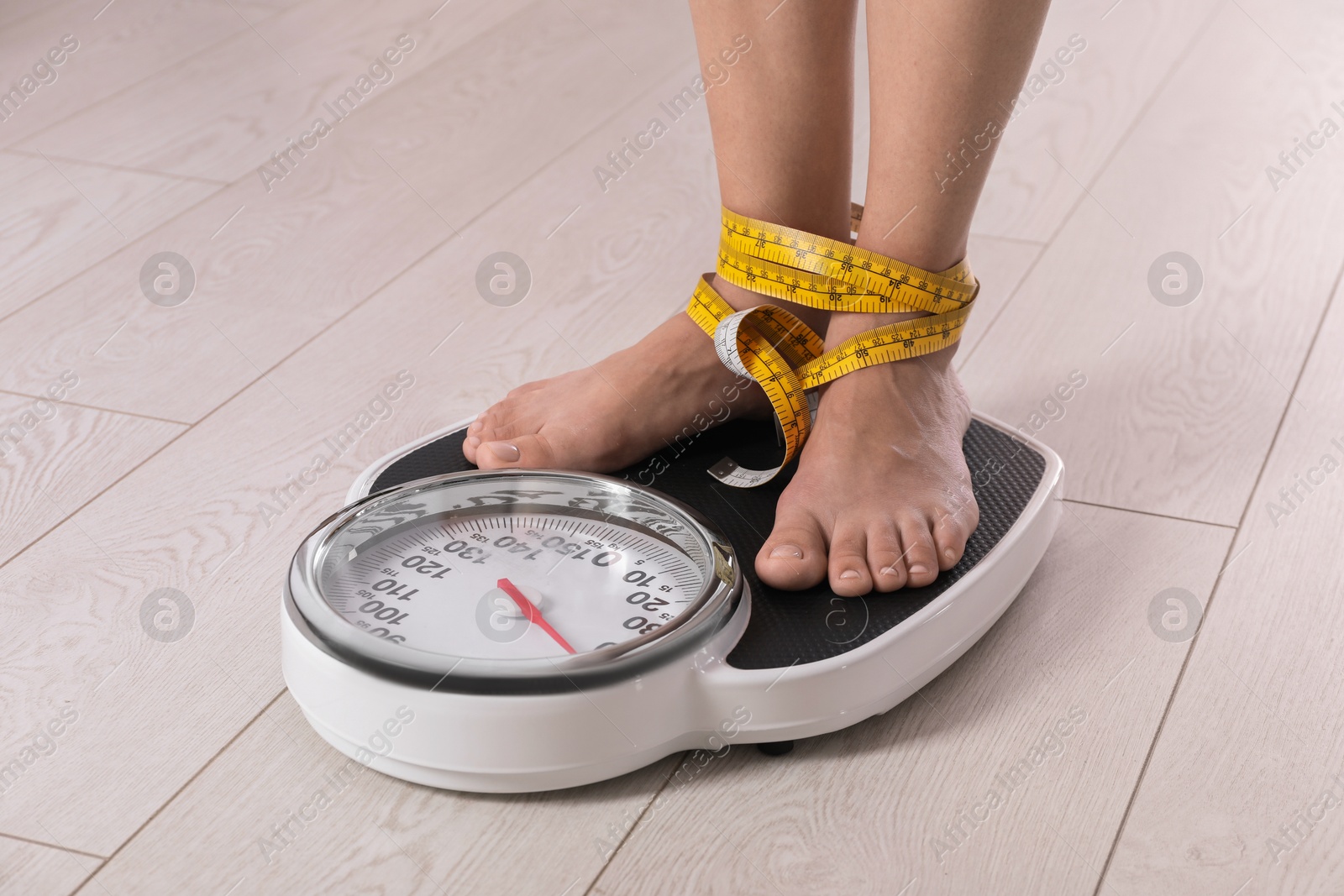 Photo of Eating disorder. Woman tied with measuring tape standing on floor scale indoors, closeup