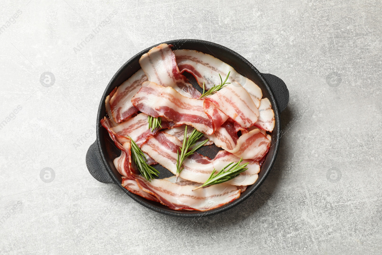 Photo of Delicious bacon slices and fresh rosemary in frying pan on light grey table, top view