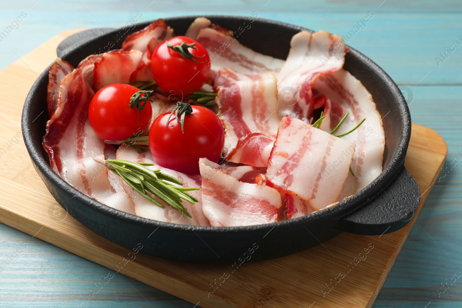 Photo of Delicious bacon slices, spices and cherry tomatoes in frying pan on light blue wooden table