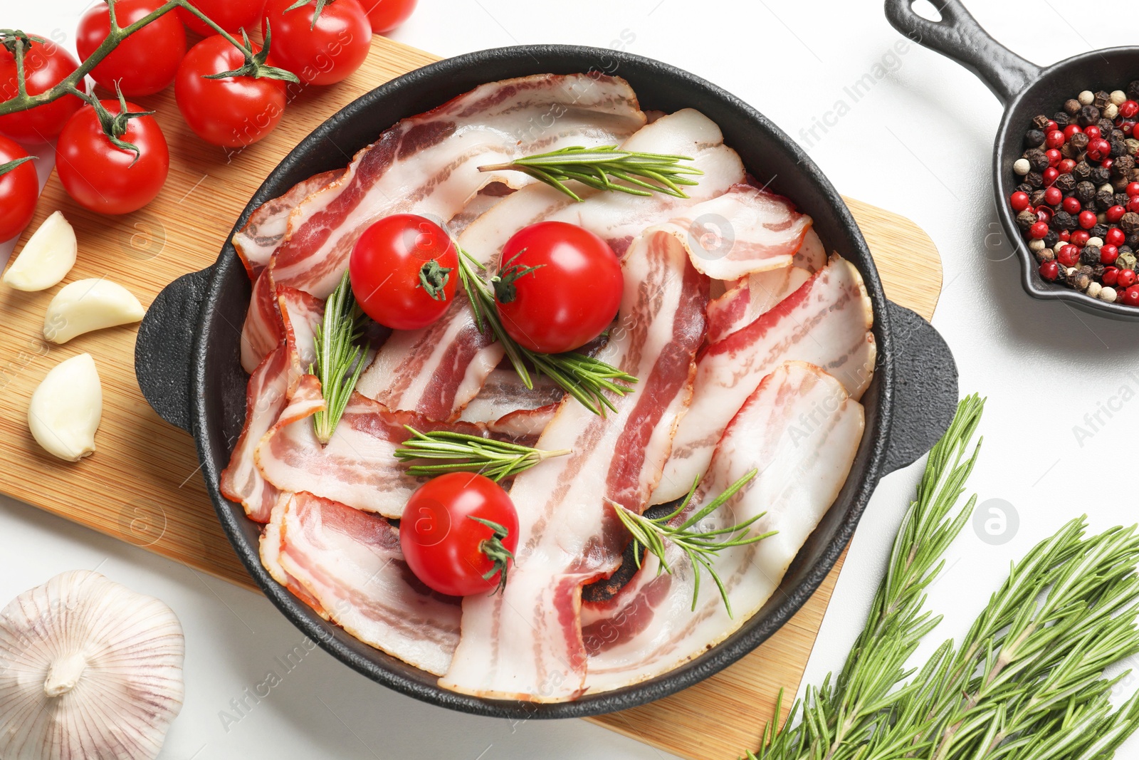 Photo of Delicious bacon slices in frying pan, spices and tomatoes on white table, flat lay