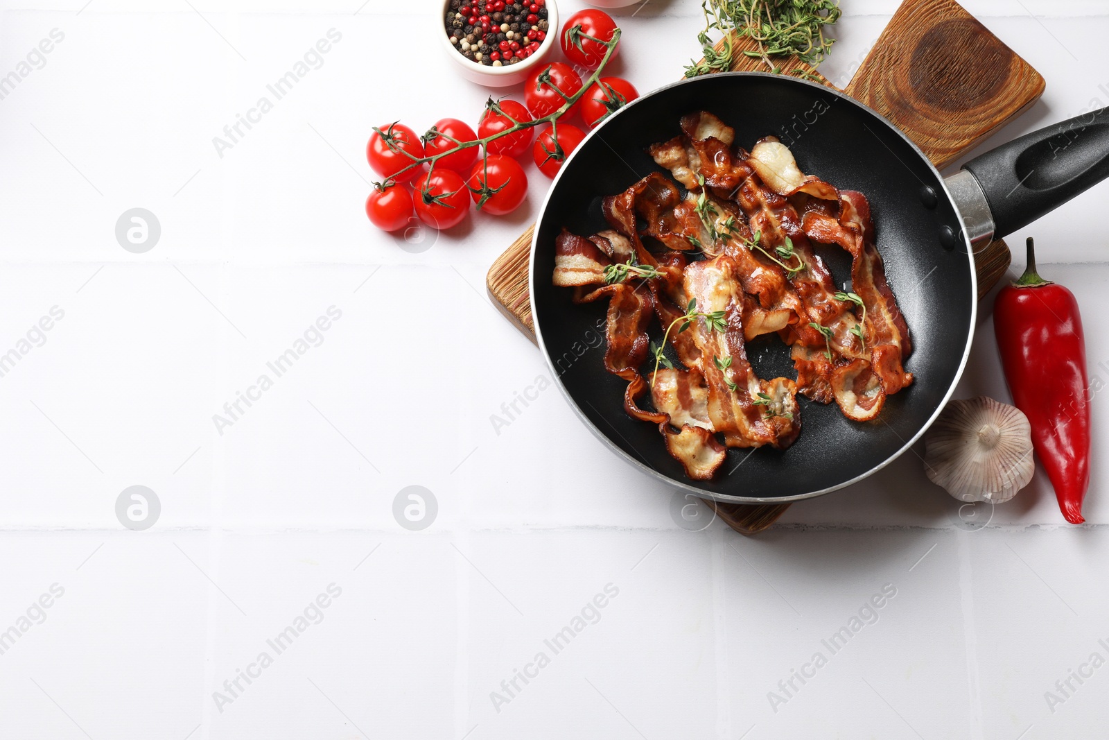 Photo of Delicious bacon slices in frying pan and products on white tiled table, flat lay. Space for text