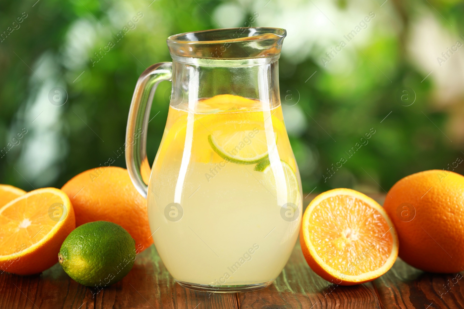 Photo of Refreshing lemonade with citruses in jug among fruits on wooden table against blurred green background