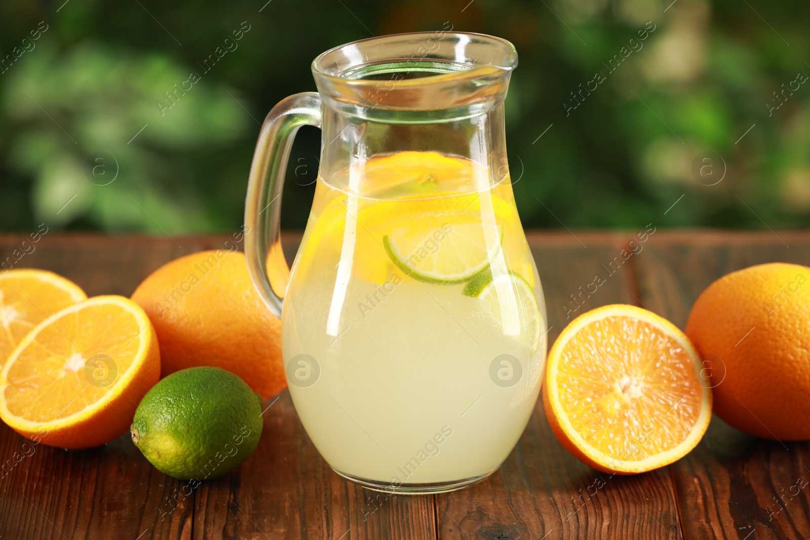 Photo of Refreshing lemonade with citruses in jug among fruits on wooden table against blurred green background